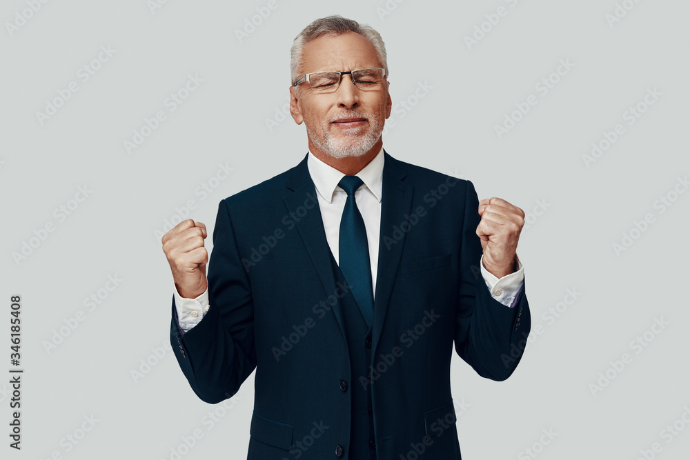 Handsome senior man in full suit gesturing and cheering while standing against grey background Handsome senior man in full suit gesturing and cheering while standing against grey background
