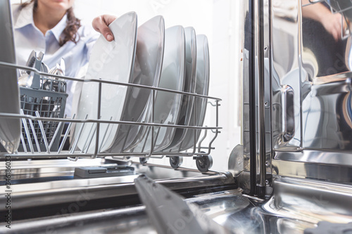 Happy young Caucasian woman, removing or placing the dishes in the dishwasher in the kitchen of her apartment. Housework concept.