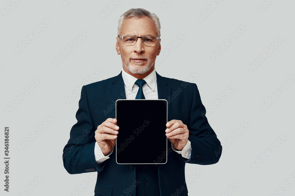 Handsome senior man in full suit pointing copy space on digital tablet and smiling while standing against grey background
