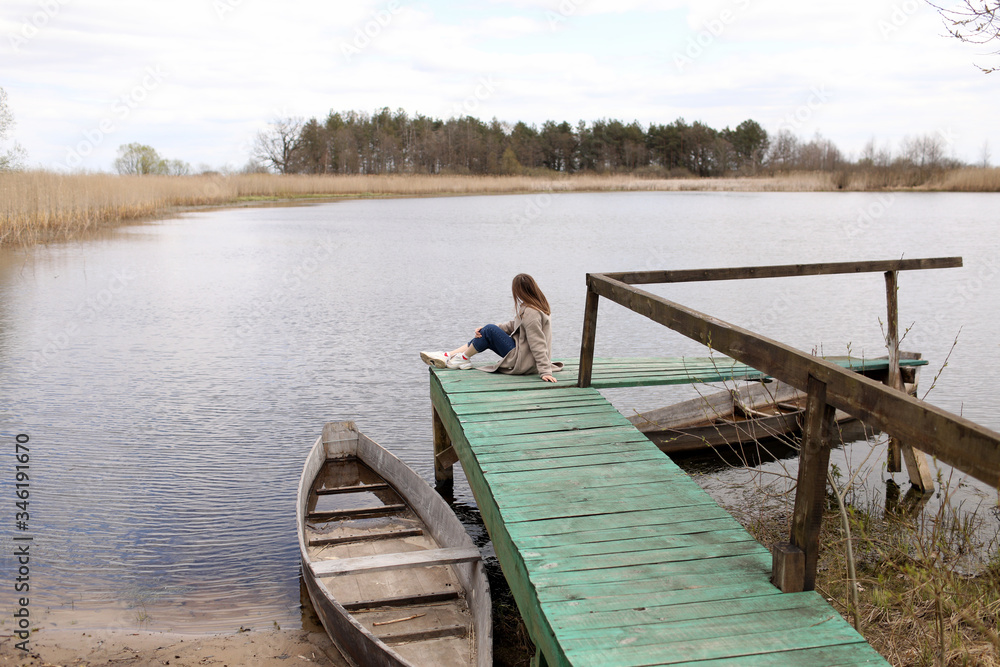 Naklejka premium Young beautiful woman sits on the wooden bridge on the river at spring day. old boat near the shore. selective focus.