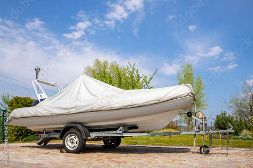 Big modern inflatable motorboat ship covered with grey or white protection tarp standing on steel semi trailer at home backyard on bright sunny day with blue sky on background. Boat vessel storage