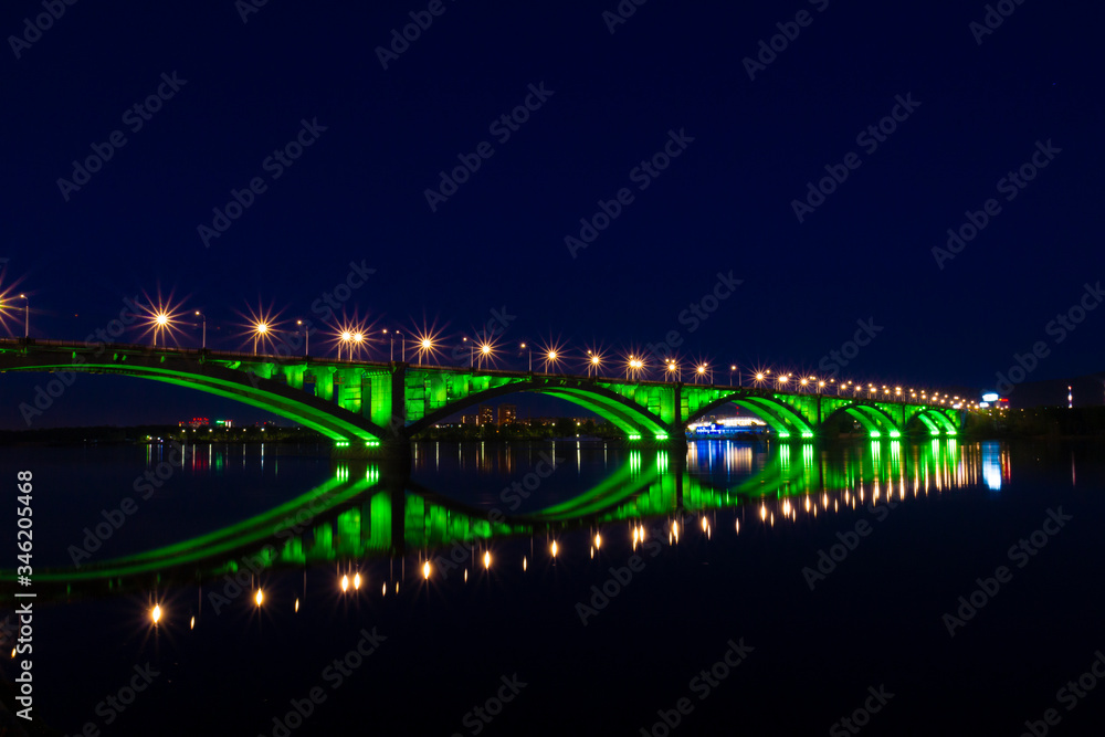 Communal bridge over the Yenisei River in Krasnoyarsk, Russia. Reflection of a bridge and illuminations in the water. Night lights of the city of Krasnoyarsk. Night urban landscape. City Lights