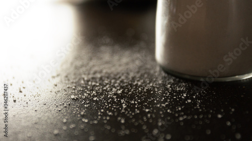 фотографія blurred photo Salt crumbled on a table near a salt shaker, white salt on a black table near a glass jar for storing spices