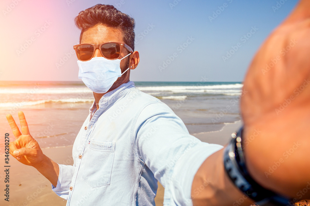 Young african american man in blue shirt and mask making a selfie portrait on the front camera of a smartphone on the ocean beach - obrazy, fototapety, plakaty 