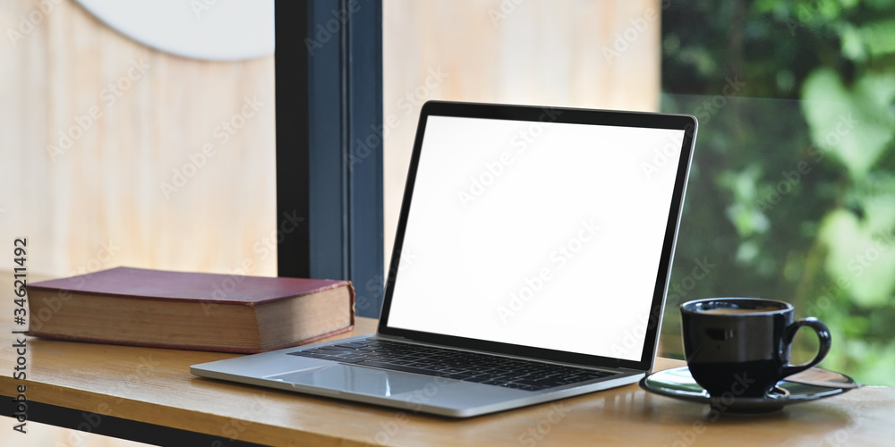 Photo of computer laptop with white blank screen, book and coffee cup putting on wooden counter bar over comfortable living room glass wall as background. Photo of computer laptop with white blank screen, book and coffee cup putting on wooden counter bar over comfortable living room glass wall as background.