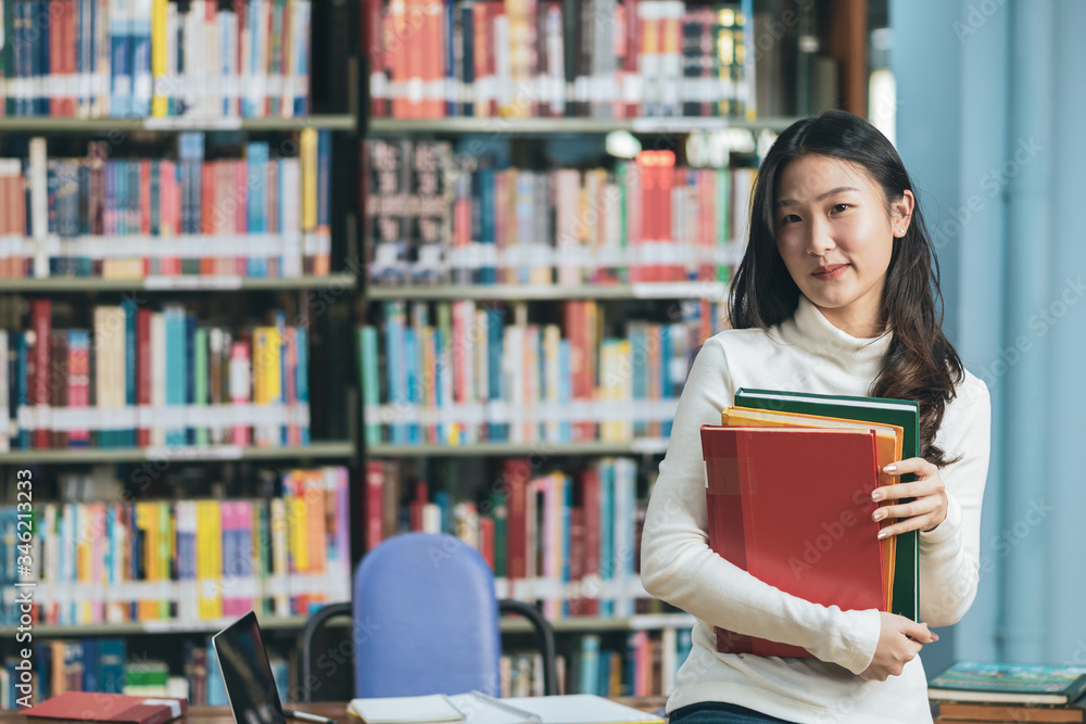 Asian young student in white casual suit reading the book in library of university or colleage.Standing and reading on a wooden table and bookcase in the background.Back to school Asian young student in white casual suit reading the book in library of university or colleage.Standing and reading on a wooden table and bookcase in the background.Back to school