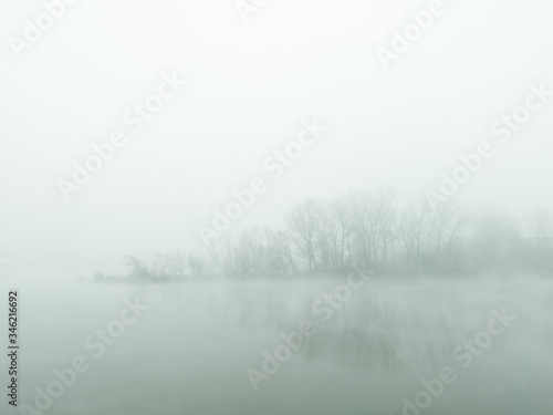 Mysterious and ghostly river island obscured by dense fog which float above flat water surface and slips through between trees. The gloomy silent autumn landscape of the river island in the thick fog.