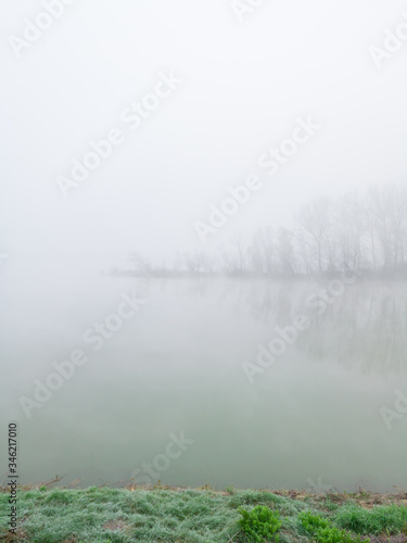 The gloomy landscape of the morning mist in the river valley. A thick fog hovers over the flat surface of the river and creeps among the trees in the woods on a barely visible river island.