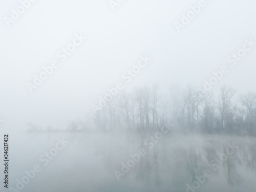 Peaceful autumn scene of mystical river island with a deciduous grove in deep fog early in morning. Calm and silent nature scene covered with thick fog. © slobodan