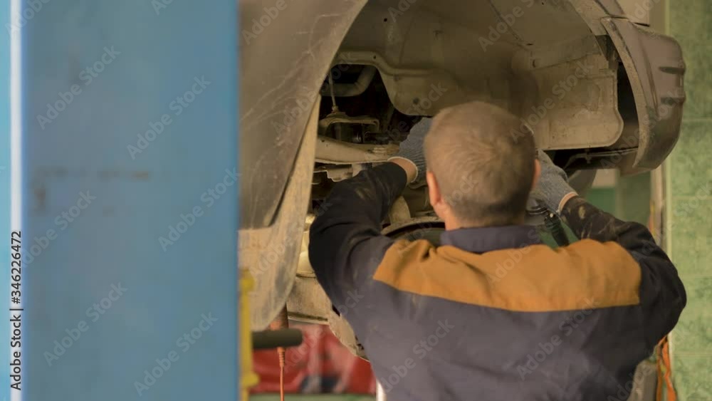 An Asian man is repairing the chassis of an old car lifted on a lift in a workshop, close-up. An Asian man is repairing the chassis of an old car lifted on a lift in a workshop, close-up.