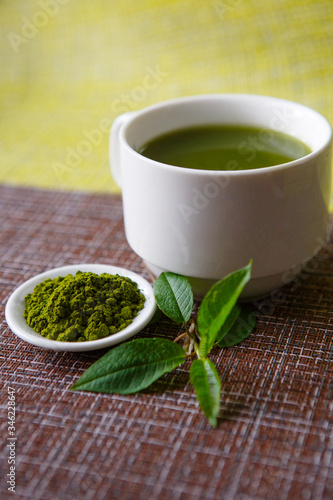 Japanese matcha green tea is poured into a white mug and on a white saucer in powder. Tea set on a textured napkin of natural flowers, decorated with a branch of green leaves. Background with space fo