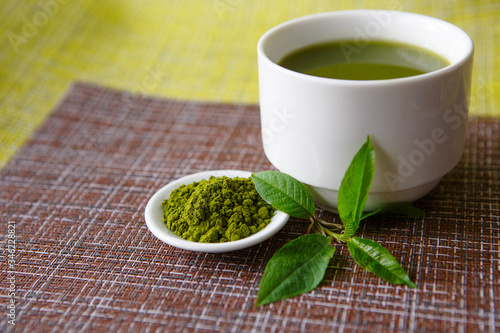 Japanese matcha green tea is poured into a white mug and on a white saucer in powder. Tea set on a textured napkin of natural flowers, decorated with a branch of green leaves. Background with space fo