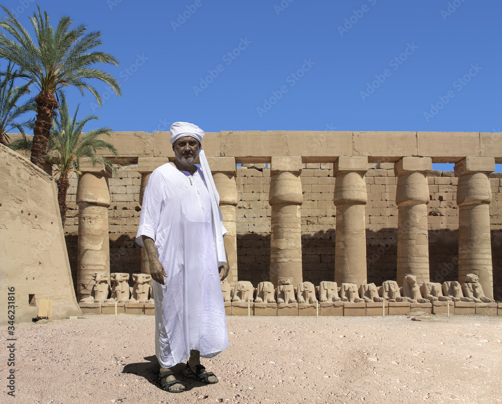 An ancient Egyptian stands in front of historical columns in Karnak in Egypt. The man in a turban is wearing a white robe and is standing in old sandals. On the top left is a palm tree.