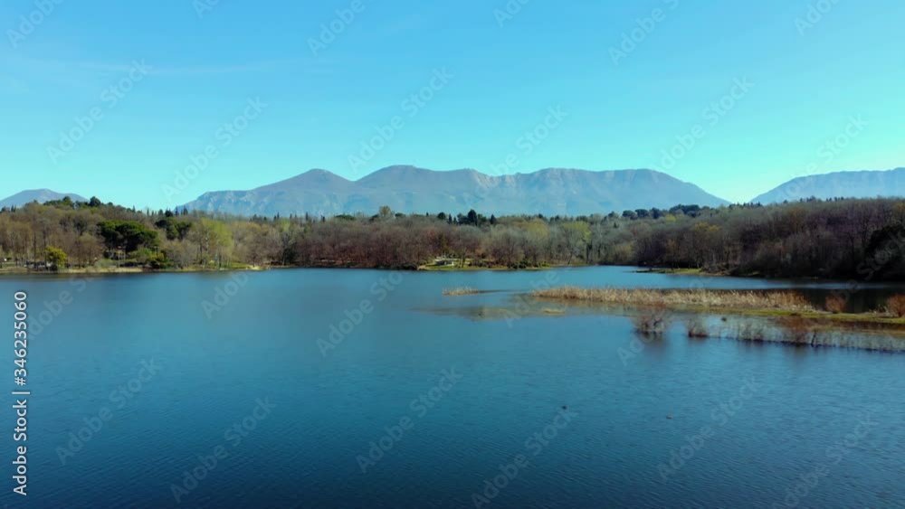 Peaceful city park with trees forest and mountain background, washed by calm clear water of lake reflecting blue sky Peaceful city park with trees forest and mountain background, washed by calm clear water of lake reflecting blue sky