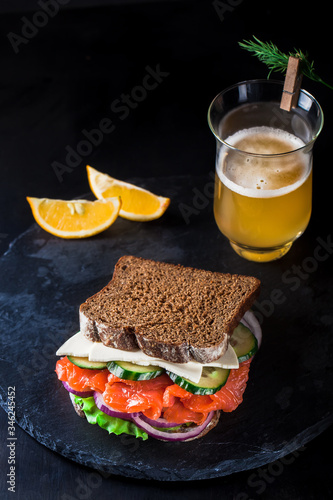 Sandwich with salmon, a glass of beer, herbs, lemon on a dark metallic background. Fish appetizer on a black table.