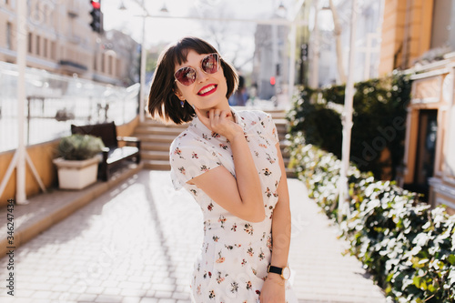 Glad young lady in white dress smiling during outdoor summer photoshoot. Short-haired brunette girl in sunglasses ejoying spring. photo