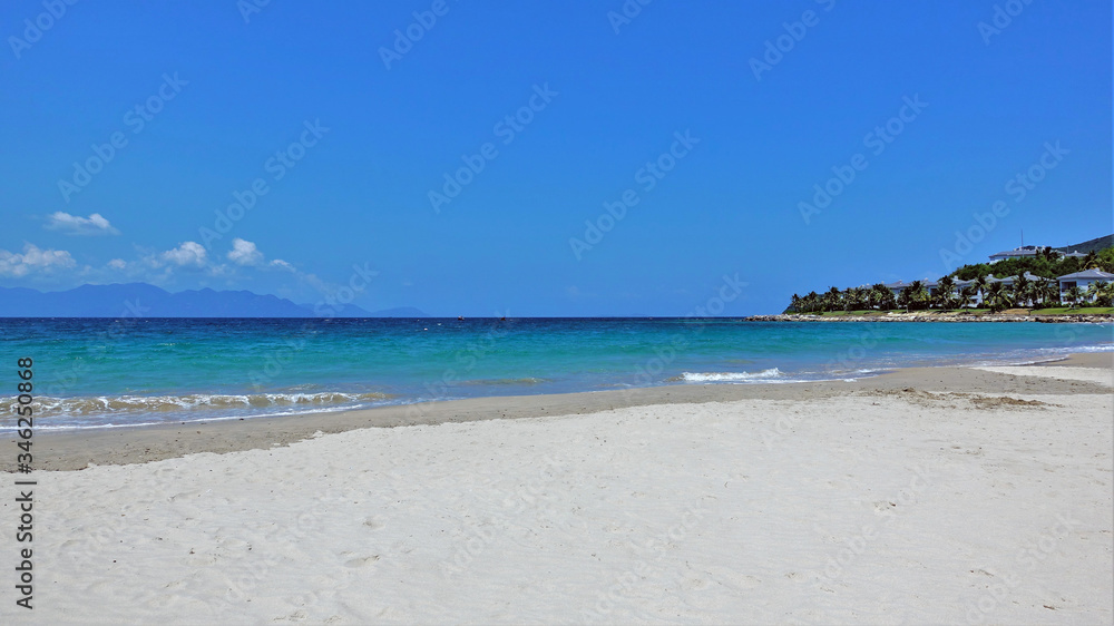 Seascape. Vietnam. Clean sand, small waves. Shades of sea water are turquoise, dark blue. Blue sky. In the distance are the silhouettes of mountains and clouds. On the coast of palm trees and villas.
