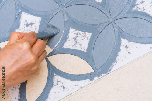 Stay at home and home improvement concept: Close up and top view of a hand holding a brush is painting a decorative template on the floor tiles into gray by using a vintage pattern stencil photo