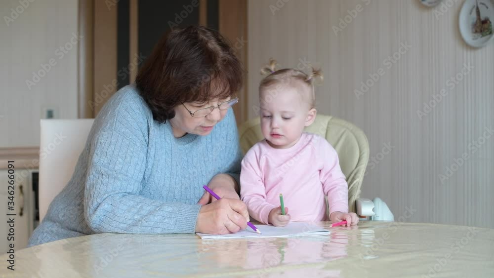 A little girl watches a nanny draw in an album. A child learns to draw with her grandmother A little girl watches a nanny draw in an album. A child learns to draw with her grandmother