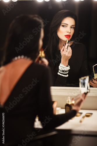 Jewelry and beauty concept. Beautiful brunette lady portrait in modern gems and black dress in studio. Woman sits by the makeup mirror and puts on red lipstick