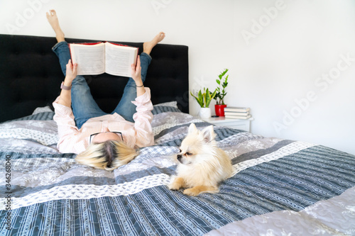 Bild auf Leinwand Blonde hair woman lying legs up on cozy bed with dog, holding open book and reading