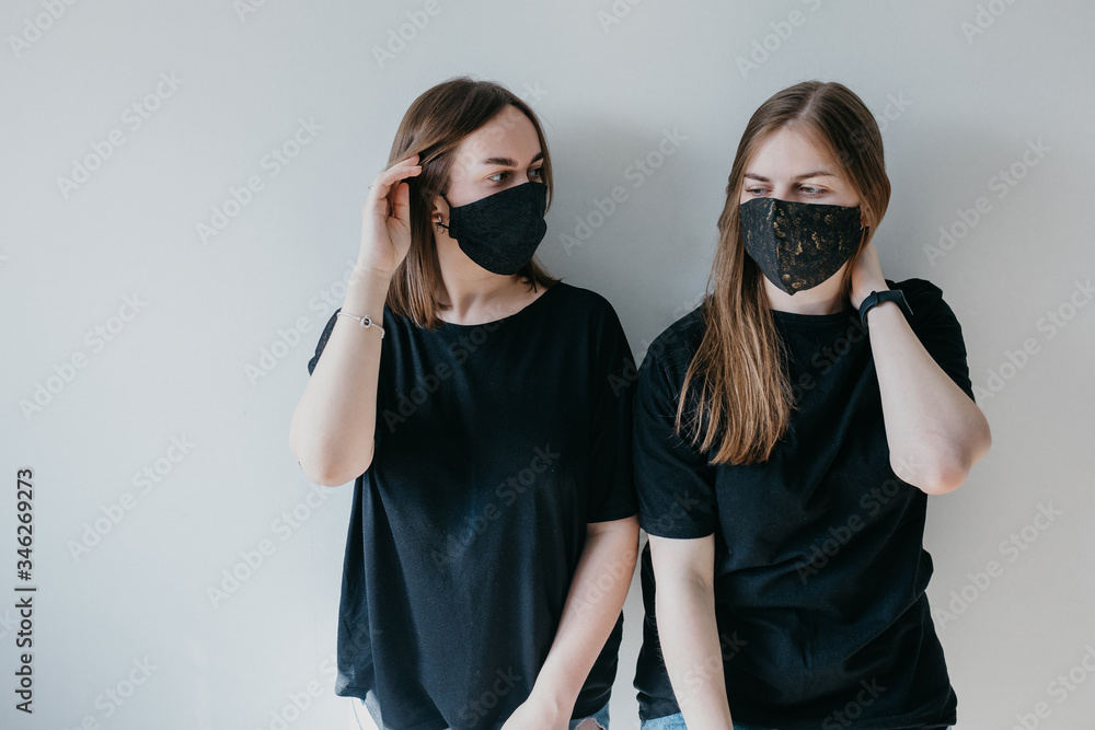 Two girls in black t-shirts and black protective masks standing near white wall Two girls in black t-shirts and black protective masks standing near white wall