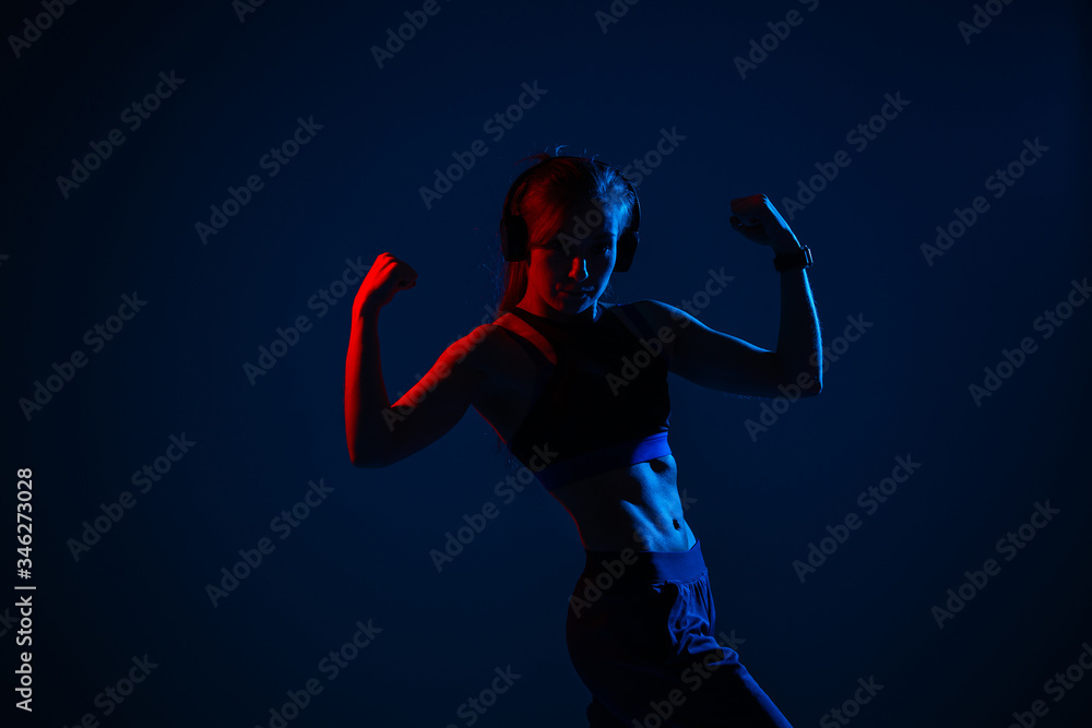 A girl with illuminated relief abdominal muscles shows biceps. Sportswoman in headphones in a neon red and blue light.