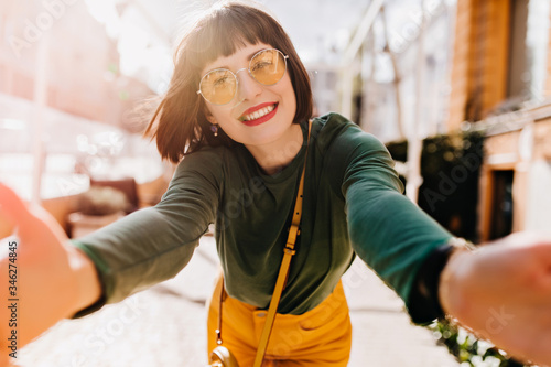 Enchanting girl with stylish haircut making selfie and laughing. Outdoor portrait of beautiful european woman wears green sweater in spring day.