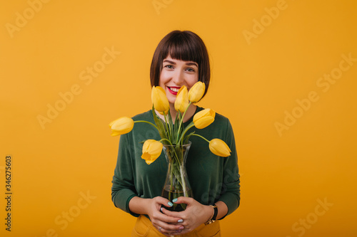 Εκτύπωση καμβά Indoor portrait of dark-haired girl in green sweater holding vase of beautiful flowers