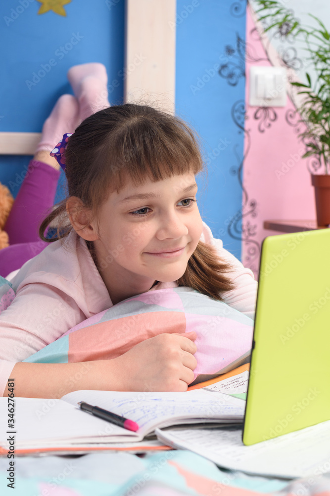 The girl is remotely studying at home, lying in bed and watching a video lesson The girl is remotely studying at home, lying in bed and watching a video lesson
