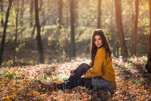 Young beautiful caucasian woman sits on the ground in a autumn park among high trees. Mysterious and seductive look of a beautiful woman