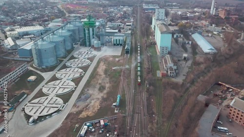 Aerial above follow from the beginning view of freight train with many carriages, moving along railway in industrial zone between granary and grain dryers. Deliver food and wheat for processing. photo