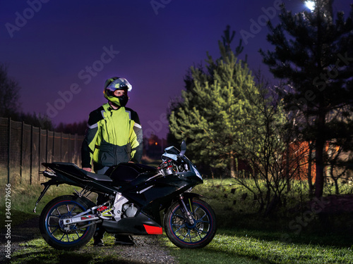 A sports bike is on the road.A young man stands nearby.In a helmet. On one side is a chain-link fence and on the other are trees. Sunset. The sky is blue and purple. Motorcycle