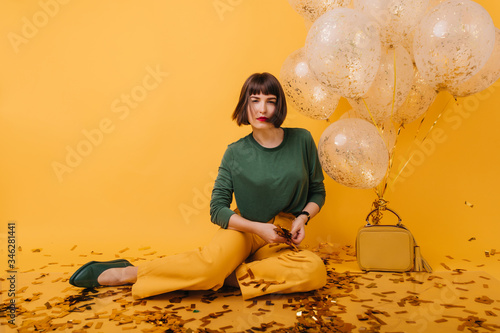 Interested woman with short hair posing after party. Magnificent dark-haired girl with serious face expression sitting on yellow background surrounded by birthday balloons.