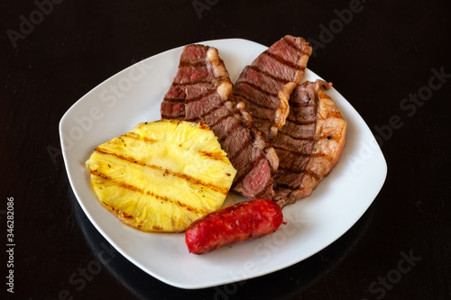 The pieces of meat are juicy, chopped, with pineapple and sausage on a white plate. Dark black background. Close-up.