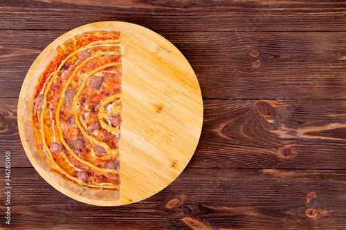 Half of salsiccia pizza with beef sausages, mozzarella, various sauces and marinated red onions on a round wooden platter which is on a wood rustic table, top view and copy space