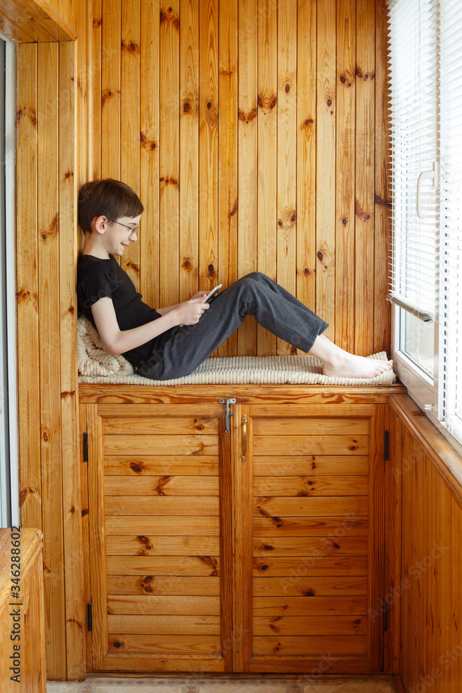 A boy teenager playing at his tablet sitting near window on a balcony. Distance learning. Wearing casual outfit. Internet technology in schoolboy's life. Gamer child. Modern generation kid. Stay home A boy teenager playing at his tablet sitting near window on a balcony. Distance learning. Wearing casual outfit. Internet technology in schoolboy's life. Gamer child. Modern generation kid. Stay home