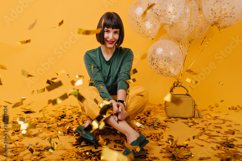 Fototapeta Naklejka Na Ścianę i Meble -  Indoor portrait of pleasant brunette girl sitting on confetti. Studio shot of cheerful european lady in green sweater posing on yellow background.