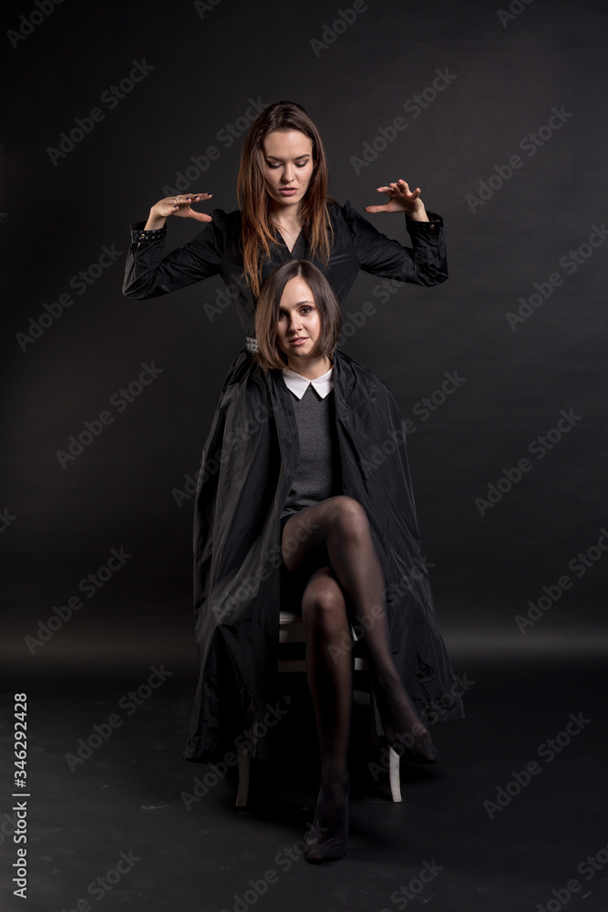 Two brunette girls in dark clothes in a photo studio on a dark background. One is sitting on a chair, the other is standing holding her hands above her. Vertical orientation.