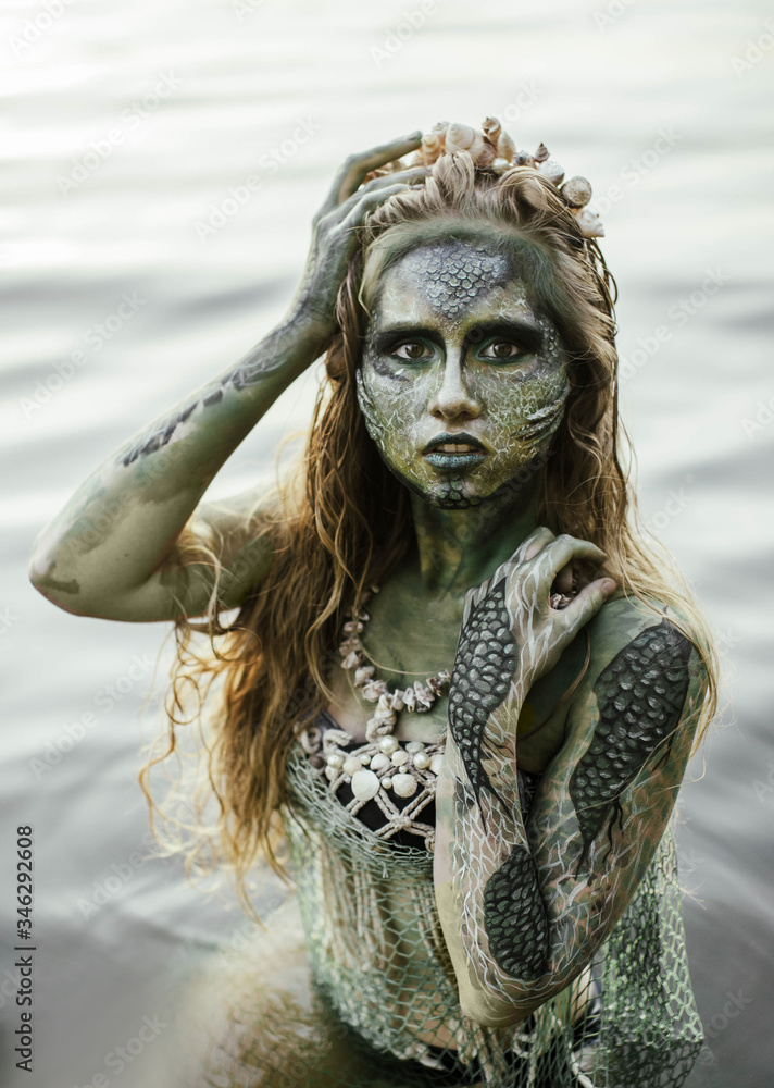 girl mermaid amphibian with green skin with body art with a necklace and a seashell swimsuit around her neck holding her hand to her serious face in the river summer in the water in the lake girl mermaid amphibian with green skin with body art with a necklace and a seashell swimsuit around her neck holding her hand to her serious face in the river summer in the water in the lake