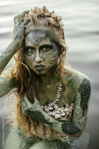 girl mermaid amphibian with green skin with body art with a necklace and a seashell swimsuit around her neck holding her hand to her serious face in the river summer in the water in the lake