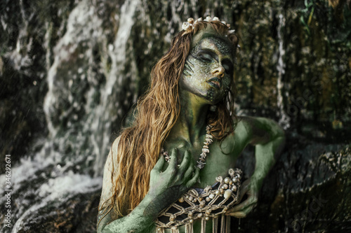 girl mermaid amphibian with green skin with body art with a necklace and a seashell swimsuit around her neck holding her hand to her serious face in the river summer in the water in the lake