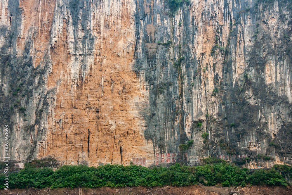 Baidicheng, China - May 7, 2010: Qutang Gorge on Yangtze River. Closeup of Straigth down brown cliff with some green foliage and darker stalactites above browm dirt shoreline..