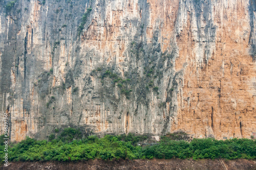 Baidicheng, China - May 7, 2010: Qutang Gorge on Yangtze River. Closeup of Straigth down brown cliff with some green foliage and darker stalactites above browm dirt shoreline..