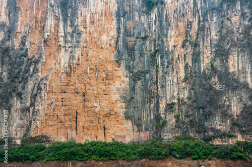 Baidicheng, China - May 7, 2010: Qutang Gorge on Yangtze River. Closeup of Straigth down brown cliff with some green foliage and darker stalactites above browm dirt shoreline..