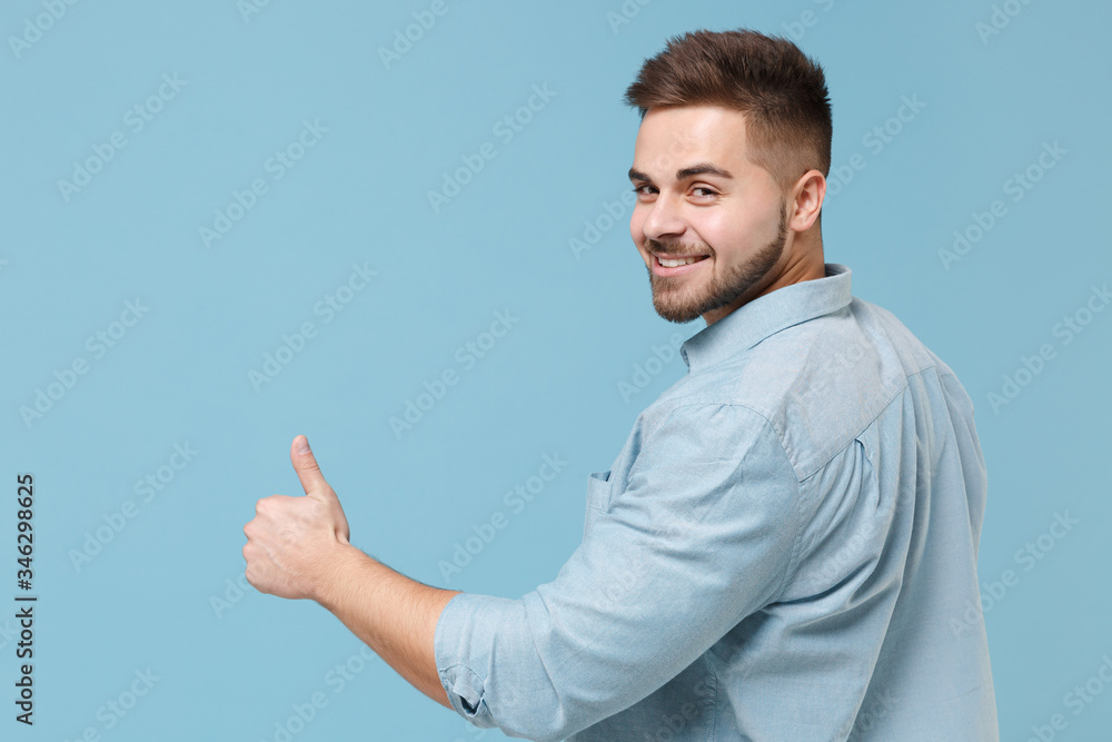 Back rear view of smiling bearded young guy 20s in casual shirt posing isolated on pastel blue background in studio. People lifestyle concept. Mock up copy space. Showing thumbs up, looking camera. Back rear view of smiling bearded young guy 20s in casual shirt posing isolated on pastel blue background in studio. People lifestyle concept. Mock up copy space. Showing thumbs up, looking camera.