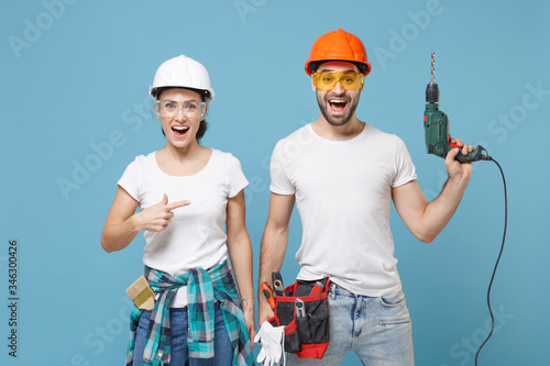 Excited couple woman man in protective helmet hardhat pointing index finger on electric drill isolated on blue background. Instruments accessories for renovation apartment room. Repair home concept. photo