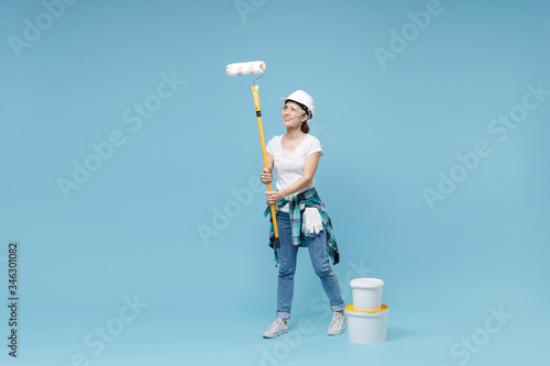 Smiling woman in protective helmet hardhat, glasses hold paint bucket, roller for wall painting isolated on blue background. Instruments accessories for renovation apartment room. Repair home concept.
