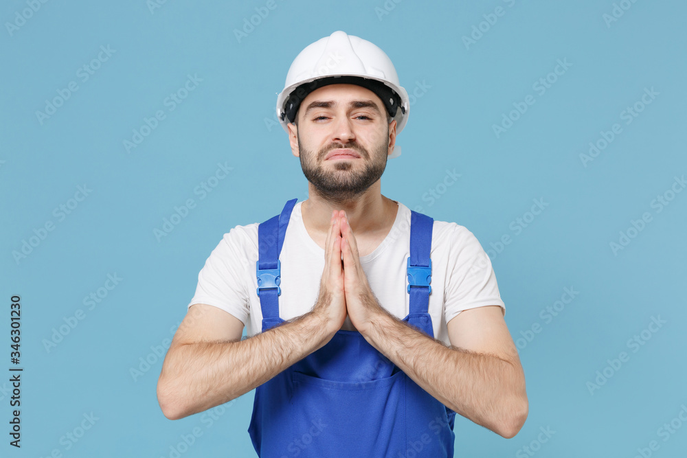 Pleading young man in coveralls protective helmet hardhat isolated on blue wall background. Instruments accessories for renovation apartment room. Repair home concept. Holding hands folded in prayer.