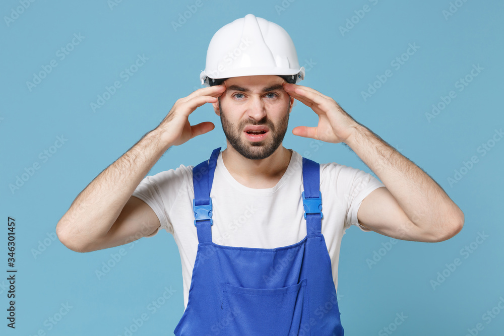 Dissatisfied young man in coveralls protective helmet hardhat isolated on blue background. Instruments accessories for renovation apartment room. Repair home concept. Put hand on head having headache.